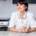 A cheerful woman leaning on a granite countertop in a modern kitchen.