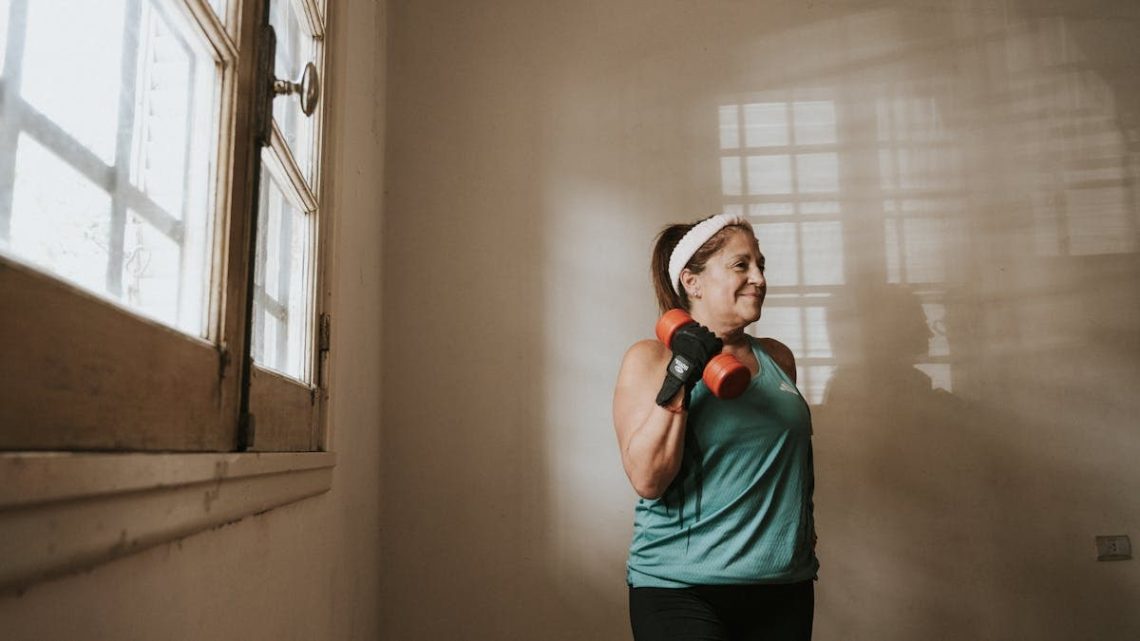Senior woman in a brightly lit room exercises with dumbbells, promoting fitness at home.