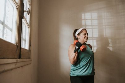 Senior woman in a brightly lit room exercises with dumbbells, promoting fitness at home.